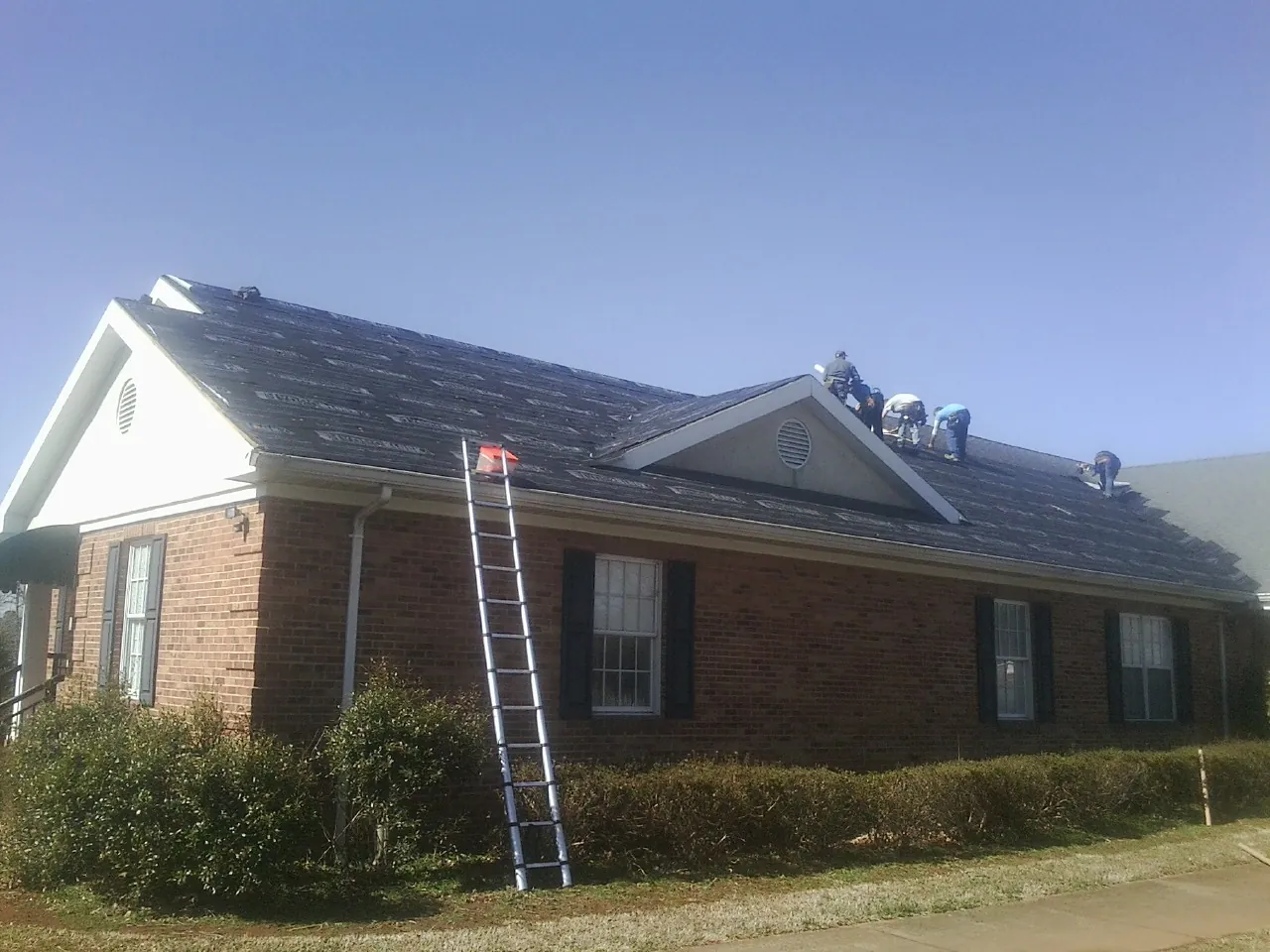 Crew tearing shingles off a brick ranch roof with a ladder set — Metal Roof Repair work by our crew in Homeland